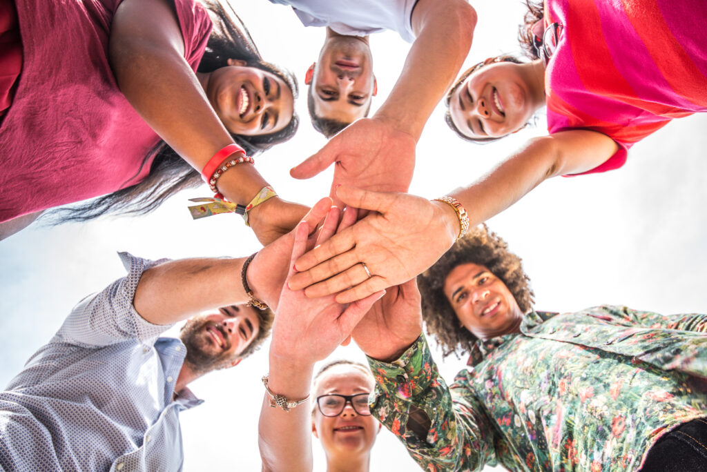 A circle of diverse people smile, put their hands in to the centre of the image. The photo is taken from the ground, looking up.