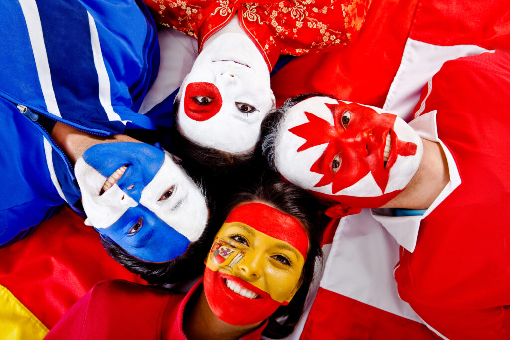 Four smiling people lay on the floor, heads together, looking up at the camera. They have different national flags painted on their faces.
