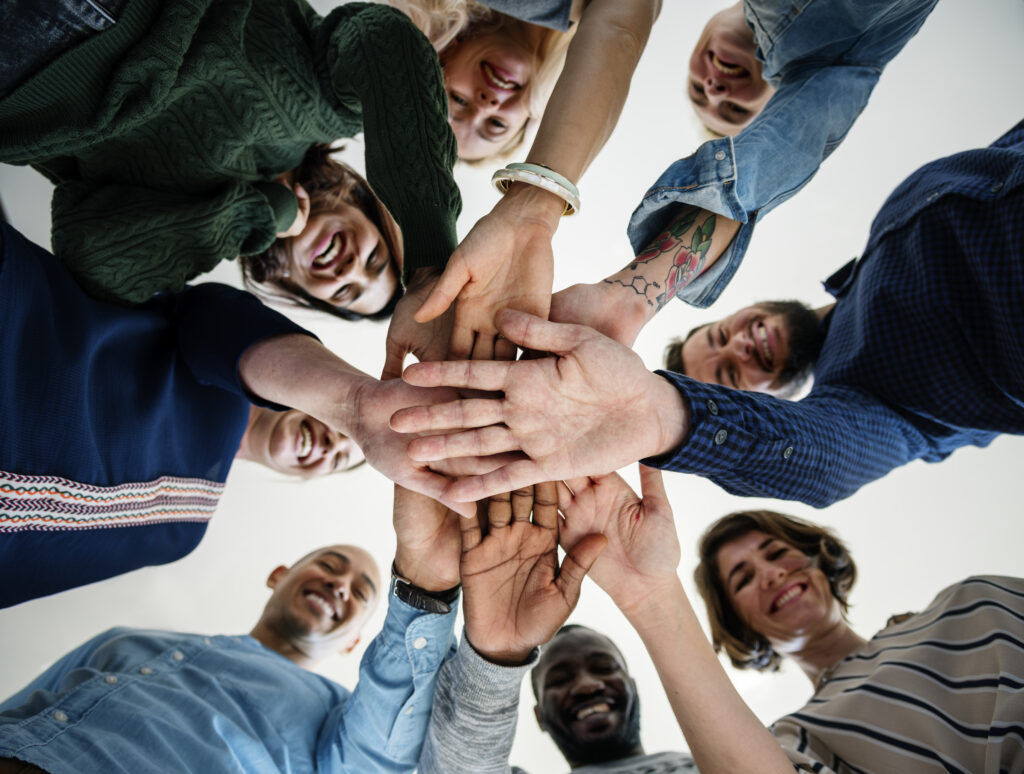 A circle of diverse people smile, put their hands in to the centre of the image. The photo is taken from the ground, looking up.