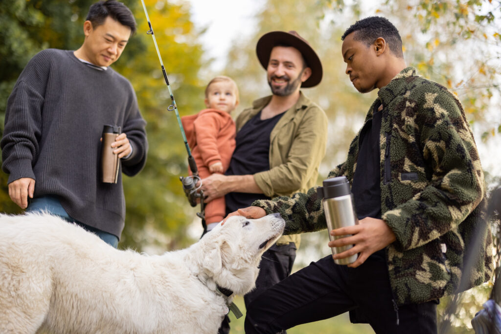 Multiracial male friends talking and having fun at picnic in nature at autumn. Concept of leisure, weekend and entertaiment. Idea of male friendship. Caucasian man with fishing rod and little son