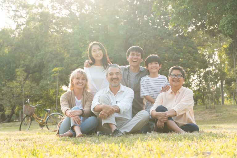 A diverse family of six people are sitting outside on a sunny day, smiling at the camera