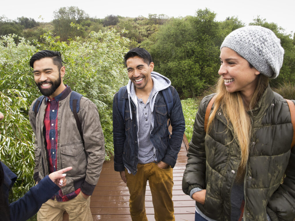 Three smiling individuals are outside on a boardwalk walking together, there is green forest in the backgroun