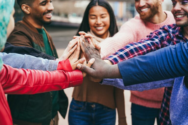 A diverse group of seven people all put one hand in to hold each other's hand