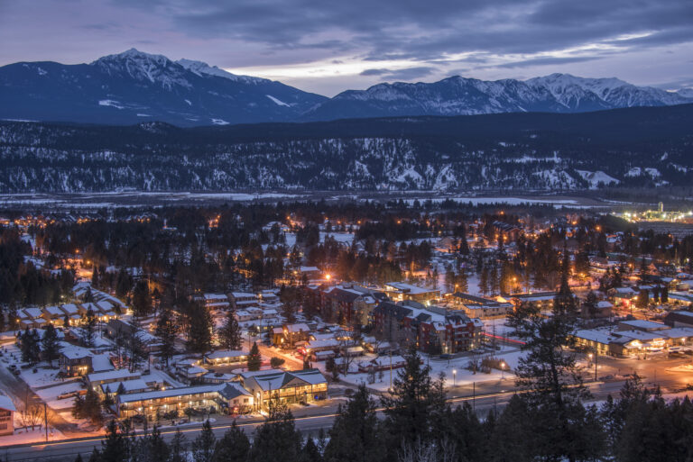 A view of the village from an above angle at dusk, street lit with orange glow against a blue mountain backdrop. Homes and buildings line the streets with conifers dotted between them, the Columbia river flows by at the back, with Purcell Mountains rising behind it.