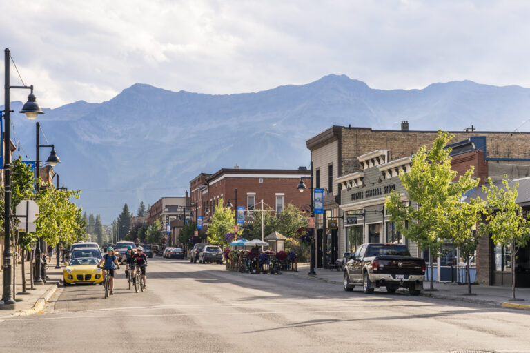 Downtown Fernie on a warm, overcast day, the street is lined with heritage storefronts, characterful lampposts and young birch trees. cyclists are approaching an intersection. Huge, hazy mountains loom in the background.