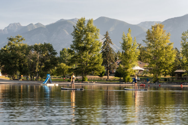 Two paddleboarders drift across Windermere Lake in front of Kinsmen Beach, which is heavily treed with conifer on a warm, hazy day. Large mountains are in the background.