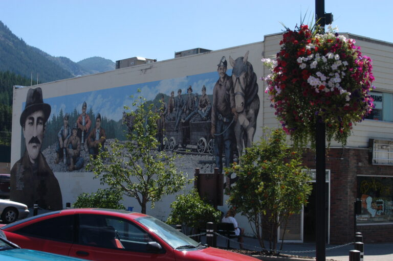 A large mural on the side of a building in downtown Sparwood depicts miners. Small trees line the street in front of the building, a lamppost hosts a large hanging basket of white and pink flowers. A mountain is visible behind the building.