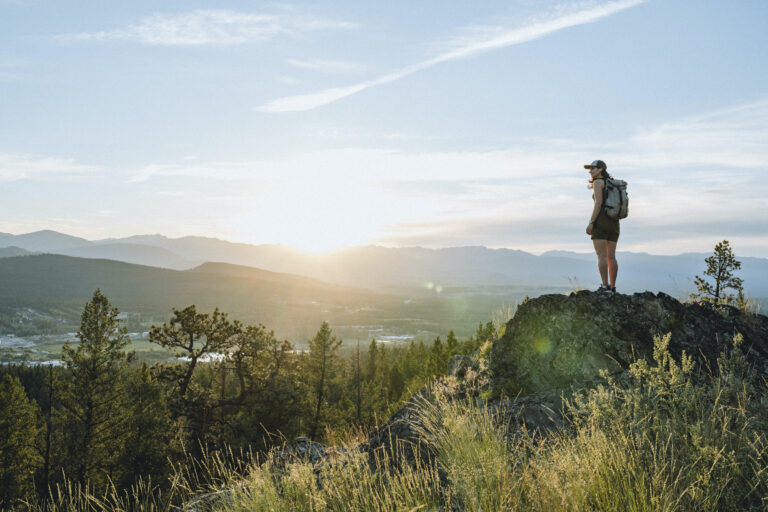 A lone hiker stands atop a rocky butte overlooking the town of Cranbrook, sat in a bowl of rock mountains. The sun on the horizon is clipped by a mountain top, creating a lens flare.